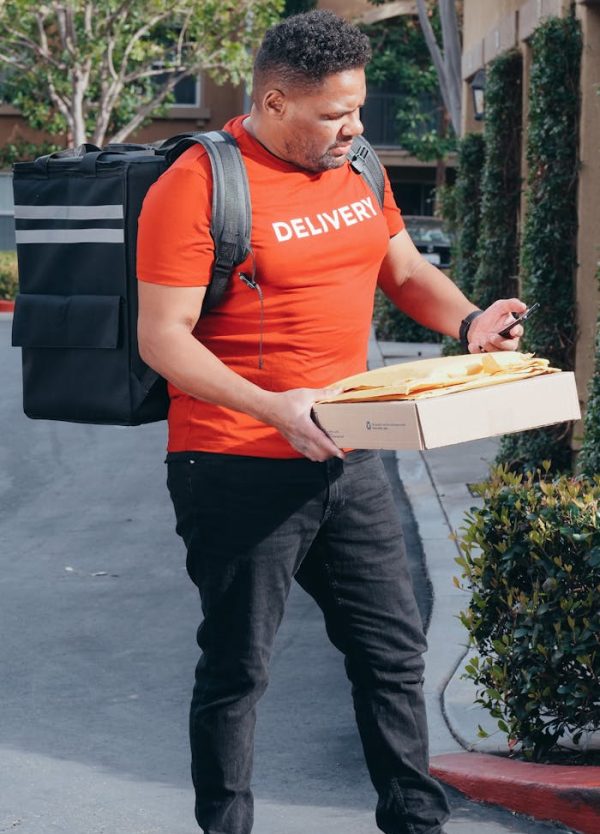 A delivery man checks his smartphone while holding a pizza box outdoors. Tall bushes and an apartment complex can be seen in the background.